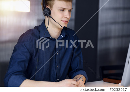 Young blond businessman using headset and computer in a darkened office, glare of light on the background. Startup business means working hard Young blond businessman using headset and computer in a darkened office, glare of light on the background. Startup business means working hard 70232917
