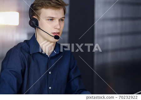 Young blond businessman using headset and computer in a darkened office, glare of light on the background. Startup business means working hard Young blond businessman using headset and computer in a darkened office, glare of light on the background. Startup business means working hard 70232922