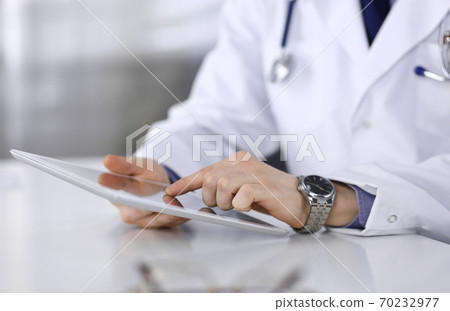 Doctor sitting and working with tablet computer in clinic at his working place, close-up. Young physician at work. Perfect medical service, medicine concept 70232977