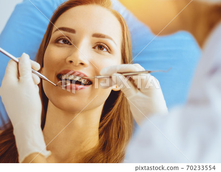 Smiling young woman with orthodontic brackets examined by dentist in sunny dental clinic. Healthy teeth and medicine concept Smiling young woman with orthodontic brackets examined by dentist in sunny dental clinic. Healthy teeth and medicine concept 70233554