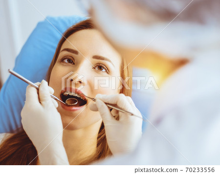 Smiling young woman with orthodontic brackets examined by dentist in sunny dental clinic. Healthy teeth and medicine concept Smiling young woman with orthodontic brackets examined by dentist in sunny dental clinic. Healthy teeth and medicine concept 70233556