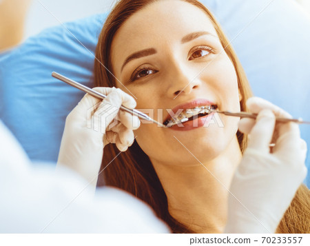 Smiling young woman with orthodontic brackets examined by dentist in sunny dental clinic. Healthy teeth and medicine concept Smiling young woman with orthodontic brackets examined by dentist in sunny dental clinic. Healthy teeth and medicine concept 70233557