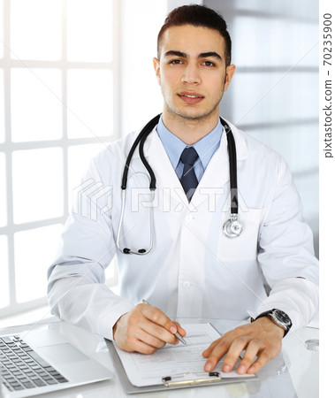 Arab doctor man is using laptop computer while filling up medication history records form sunny clinic. Medicine concept 70235900
