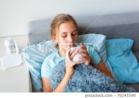 Sick teenage little girl with high fever and headache staying in bed and holding hot cup of tea Sick teenage little girl with high fever and headache staying in bed and holding hot cup of tea 70243590