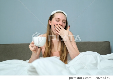 Sleepy yawning young woman in bed with cup of tea 70246139
