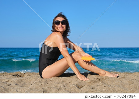 Mature beautiful woman sitting on beach using sun protection cream Mature beautiful woman sitting on beach using sun protection cream 70247084