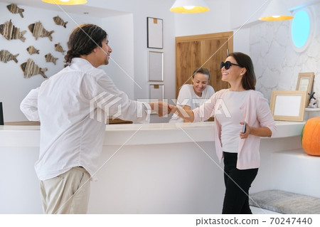 Receptionists standing at the front desk talking and greeting woman guest 70247440