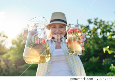Close-up of jug and glass with mint strawberry drink in hands of woman Close-up of jug and glass with mint strawberry drink in hands of woman 70247472