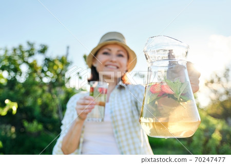 Close-up of jug and glass with mint strawberry drink in hands of woman Close-up of jug and glass with mint strawberry drink in hands of woman 70247477