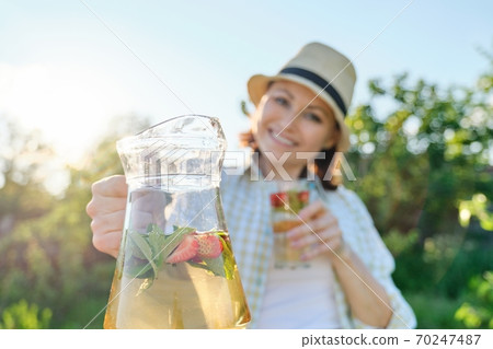 Close-up of jug and glass with mint strawberry drink in hands of woman Close-up of jug and glass with mint strawberry drink in hands of woman 70247487