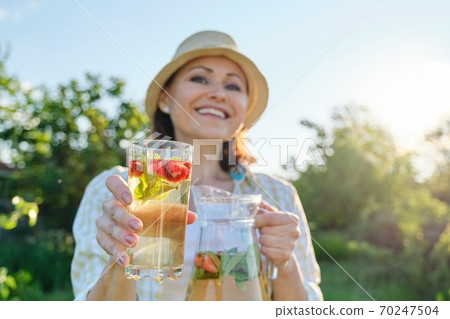 Close-up of jug and glass with mint strawberry drink in hands of woman Close-up of jug and glass with mint strawberry drink in hands of woman 70247504