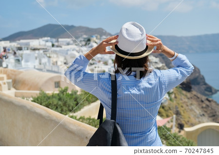 Happy woman tourist in hat with backpack looking at scenery of Santorini island Happy woman tourist in hat with backpack looking at scenery of Santorini island 70247845