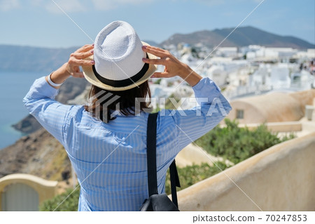 Happy woman tourist in hat with backpack looking at scenery of Santorini island Happy woman tourist in hat with backpack looking at scenery of Santorini island 70247853