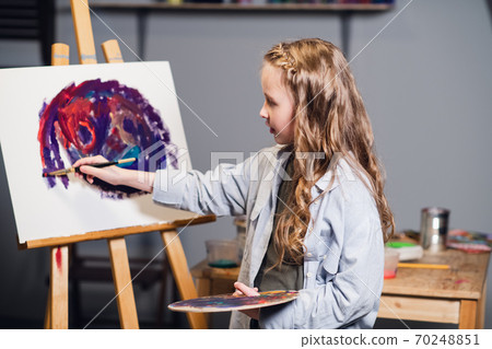 A young cute artist girl is learning to draw in an art workshop. Draws on an easel with oil paints and a brush. A young cute artist girl is learning to draw in an art workshop. Draws on an easel with oil paints and a brush. 70248851
