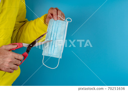 man in yellow jacket trying to cut a protective mask in half against corona virus on blue background, high alert and the battle with the disease or rebely against wearing masks 70251636