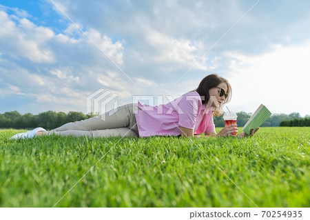 Mature woman reading book, female lying on green grass with drink Mature woman reading book, female lying on green grass with drink 70254935