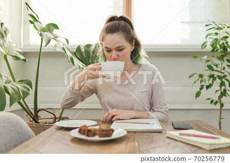 Teen girl eating cupcakes, drinking tea, sitting at table at home 70256779
