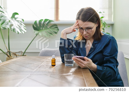 Female in pajamas sitting at home, drinking water with medicine, reading smartphone 70256810