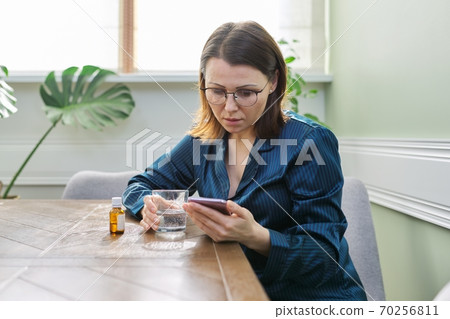 Female in pajamas sitting at home, drinking water with medicine, reading smartphone 70256811