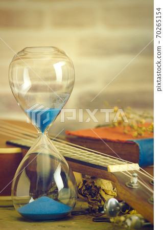 A vintage hourglass and ukulele with an old book and brass pen on a wooden table and brick background in the morning. Closeup and copy space. The concept of memories or things in the past. 70265154