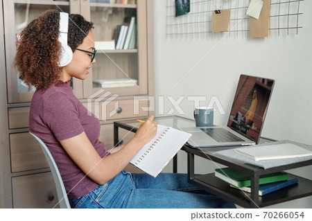 Young diligent student in casualwear and headphones making notes in copybook 70266054