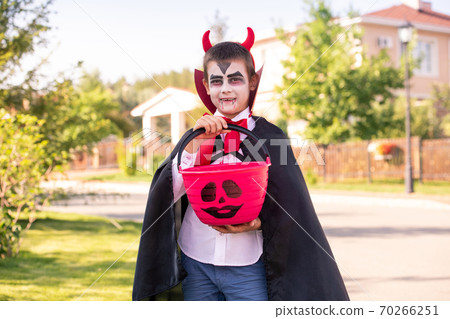 Cheerful boy in halloween costume of devil with horns on his head holding treats Cheerful boy in halloween costume of devil with horns on his head holding treats 70266251