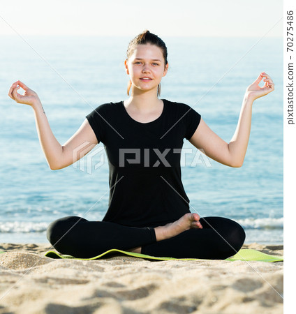 Young girl in black T-shirt is sitting and doing meditation on the beach 70275486