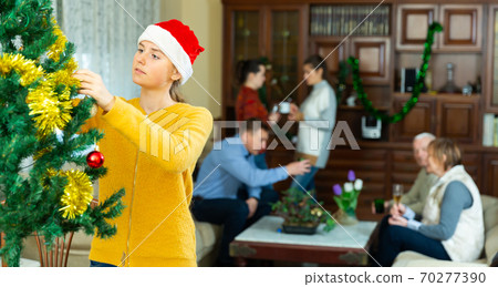 Teen girl decorates a Christmas tree. Big family has a conversation in the room Teen girl decorates a Christmas tree. Big family has a conversation in the room 70277390