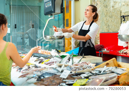 Smiling woman seller preparing raw langoustines for female buyer at seafood market, weighing order on scales 70280053