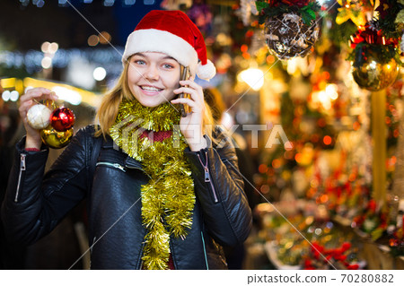 Girl in Christmas hat with phone choosing decorations at fair Girl in Christmas hat with phone choosing decorations at fair 70280882