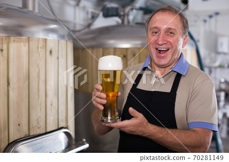 Brewer is standing with beer with foam in glass on his workplace Brewer is standing with beer with foam in glass on his workplace 70281149