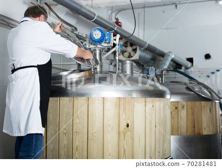 Adult brewer is standing with tube near reservoir for beer 70281481