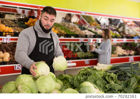 Male shop assistant lays fresh cabbage on counter in grocery shop Male shop assistant lays fresh cabbage on counter in grocery shop 70282161
