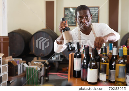 Smiling salesman in apron proposing wine in bottles in winery shop Smiling salesman in apron proposing wine in bottles in winery shop 70282935
