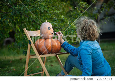 Adult woman in a blue shirt paints a pumpkin in the garden. 70283843