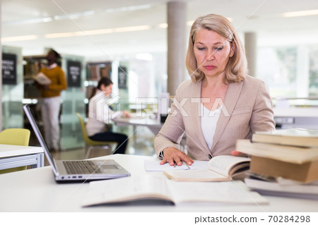Mature confident woman with laptop and book in public library Mature confident woman with laptop and book in public library 70284298