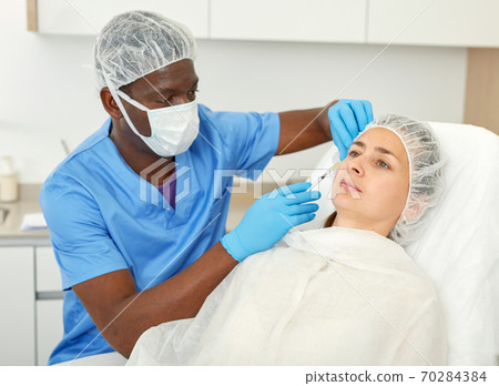 Doctor in mask is doing injection to woman patient in skin of face in estetic clinic 70284384