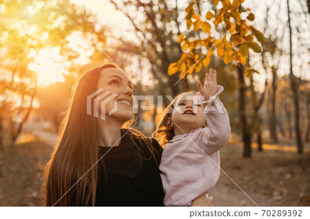 Happy family mom and toddler baby girl playing outdoors in fall park. Little girl and her mother in the autumn park Happy family mom and toddler baby girl playing outdoors in fall park. Little girl and her mother in the autumn park 70289392