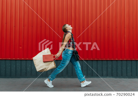 Happy girl with shopping paper bag on red wall shop background. Young woman holding shopping bag full of groceries and purchases after shopping 70290024