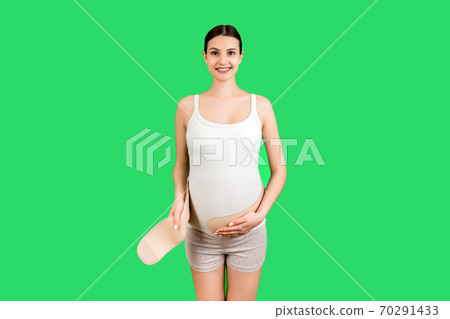Portrait of pregnant woman putting on a bandage at green background with copy space. Orthopedic abdominal support belt concept Portrait of pregnant woman putting on a bandage at green background with copy space. Orthopedic abdominal support belt concept 70291433