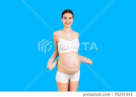 Portrait of pregnant woman in underwear putting on supporting bandage for reducing backache at blue background with copy space. Orthopedic abdominal support belt concept 70291441