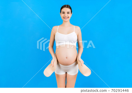 Portrait of pregnant woman in underwear dressing pregnancy corset on the third trimester at blue background with copy space. Orthopedic abdominal support belt concept 70291446