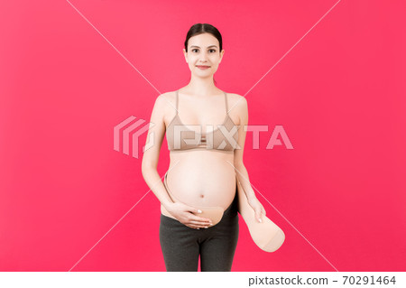Portrait of pregnant woman dressing maternity belt at pink background with copy space. Support belt against backache. Orthopedic abdominal support belt concept Portrait of pregnant woman dressing maternity belt at pink background with copy space. Support belt against backache. Orthopedic abdominal support belt concept 70291464