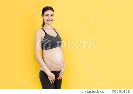 Portrait of pregnant woman wearing pregnancy bandage at yellow background with copy space. Orthopedic abdominal support belt concept Portrait of pregnant woman wearing pregnancy bandage at yellow background with copy space. Orthopedic abdominal support belt concept 70291468