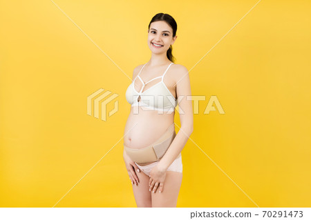 Portrait of pregnant woman in underwear wearing elastic maternity band at yellow background with copy space. Orthopedic abdominal support belt concept 70291473