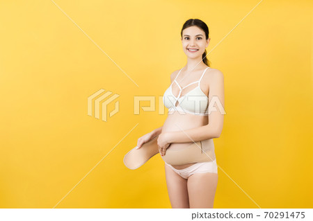 Portrait of pregnant woman in colorful underwear putting on a bandage at yellow background with copy space. Orthopedic abdominal support belt concept Portrait of pregnant woman in colorful underwear putting on a bandage at yellow background with copy space. Orthopedic abdominal support belt concept 70291475