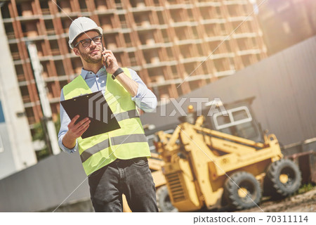 Important call. Young civil engineer or construction supervisor wearing helmet talking by phone while inspecting building site 70311114