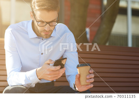 Business message. Focused businessman using smartphone and drinking coffee while sitting on the bench outdoors 70311135