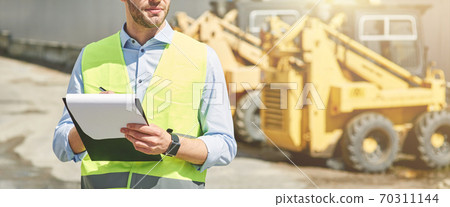 Young civil engineer wearing helmet writing report while standing at construction site Young civil engineer wearing helmet writing report while standing at construction site 70311144