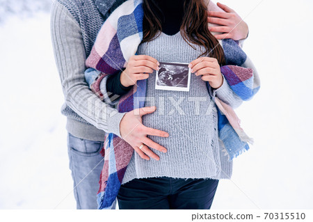 Close up man holding woman's pregnant belly and the woman holding ultrasound pregnancy picture in her hand while they stand on snowy winter park. Close up man holding woman's pregnant belly and the woman holding ultrasound pregnancy picture in her hand while they stand on snowy winter park. 70315510